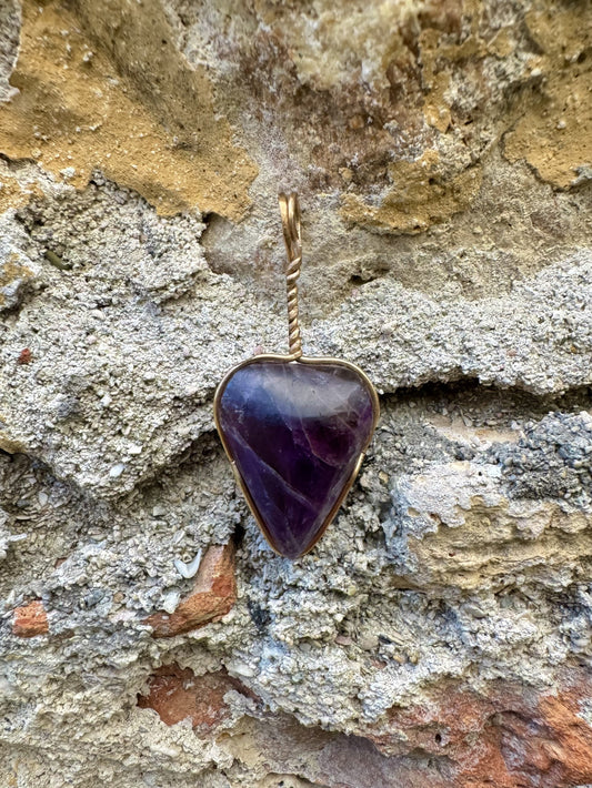 Heart-shaped purple stone pendant on a gold chain against a stone wall background