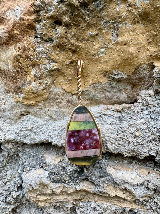 Multicolored teardrop pendant with a gold wrap against a stone wall background